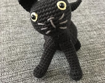 May include: A handmade black crochet cat figurine with yellow eyes and gray whiskers. The cat has pointed ears and is posed in a playful stance. The yarn is tightly woven, giving the cat a textured appearance.