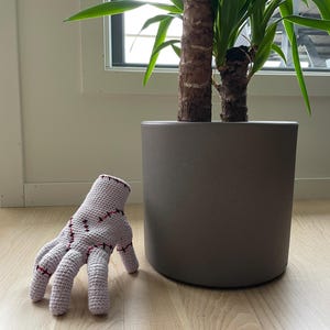 May include: A light purple, crocheted hand with red stitching details. The hand is resting on a light wood floor next to a gray plant pot with a green plant inside.