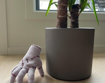 May include: A light purple, crocheted hand with red stitching details. The hand is resting on a light wood floor next to a gray plant pot with a green plant inside.