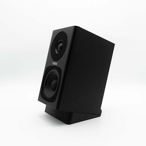 Puede incluir: Un altavoz negro con dos componentes circulares y la marca "Fostex" impresa. El altavoz está inclinado y descansa sobre una base negra, sobre un fondo blanco.