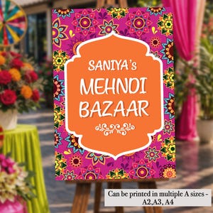 Op de afbeelding: Decoratief bord voor "Saniya's Mehndi Bazaar" met een levendig bloemenpatroon in roze, geel en oranje. Het bord wordt weergegeven op een houten ezel, met tekst in een witte, sierlijke lijst. Het bord kan in meerdere A-maten worden afgedrukt.