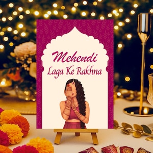 May include: A decorative sign with the text "Mehendi Laga Ke Rakhna" in a stylized font, featuring an illustration of a woman with hennaed hands. The sign is displayed on a small wooden easel, with colorful floral accents and decorative items.