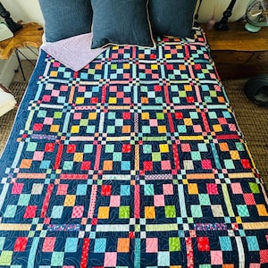 May include: A colorful patchwork quilt with a navy blue background, featuring squares of red, pink, yellow, green, and blue. The quilt is on a bed with navy blue pillows and a light purple quilted bed runner.