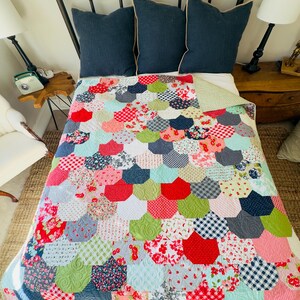 May include: A colorful handmade quilt with a scalloped pattern, featuring a variety of fabrics in red, green, blue, and white. The quilt is on a bed with navy blue pillows and a dark metal bed frame. The quilt's design includes floral, gingham, and solid patterns.