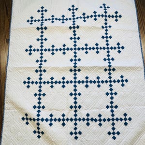 May include: A cream-colored quilt with a geometric pattern of dark blue diamond shapes. The quilt features a repeating cross-like design, with quilted diamond patterns in the background. The edges are finished with a blue border.