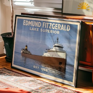 Może przedstawiać: Oprawiony wydruk statku Edmund Fitzgerald na jeziorze Superior, z tekstem "EDMUND FITZGERALD LAKE SUPERIOR EST. 1958." Statek jest brązowo-biały, na tle błękitnego nieba i wody. Wydruk jest w czarnej ramie.
