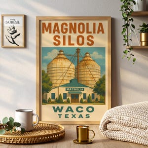 May include: A framed print with the text "MAGNOLIA SILOS WACO TEXAS" in a vintage style. The artwork shows two large silos, a building with a green roof, and trees. The print is displayed on a wooden surface with a coffee mug and candle.