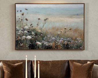 May include: A rectangular landscape painting in a brown frame featuring a field of wildflowers in muted tones of green, brown, and white. The artwork is displayed above a brown sofa with three lit taper candles and a book on a coffee table.