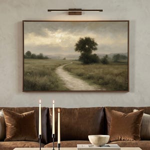 May include: A rectangular landscape painting in a brown frame depicting a dirt path winding through a grassy field toward trees under a cloudy sky. The artwork is mounted on a wall above a brown sofa with three lit taper candles on a table.