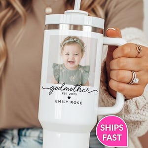 May include: White insulated tumbler with a handle, featuring a photo of a baby girl. The tumbler has the word "godmother" in script, along with "EST 2025" and "EMILY ROSE" printed below the photo. A pink sticker with "SHIPS FAST" is visible.