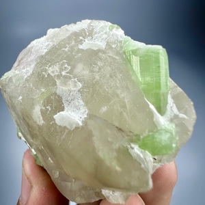 May include: A close-up of a raw, uncut mineral specimen. The stone is primarily a pale, translucent color with a crystalline structure. A vibrant green crystal is embedded within the larger stone, adding a striking contrast. The overall texture appears rough and natural.