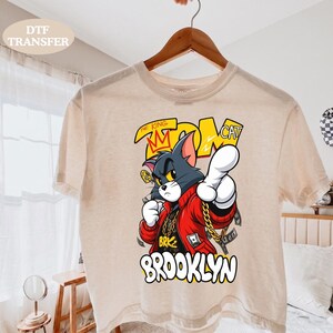 May include: A beige short-sleeve t-shirt featuring a graphic of a cartoon cat wearing a red jacket and gold chain. The text reads THE KING TOM CAT and BROOKLYN. The cat is pointing forward with one hand and has a crown above its head.
