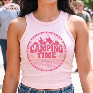May include: Light pink tank top with a circular graphic that says "Camping Time" in pink. The graphic features mountains and a winding river design. The words "DTF TRANSFER" are in the upper left corner.