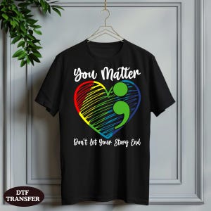 May include: A black short-sleeve t-shirt featuring a graphic of a multicolored heart with a green semicolon inside. The text "You Matter" is printed above the heart, and the text "Don't let Your Story End" is printed below the heart in white.