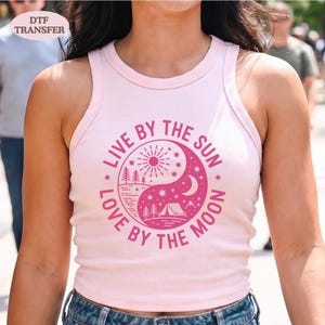 May include: A light pink tank top with a round neckline. The front features a pink yin-yang design with the words "LIVE BY THE SUN" and "LOVE BY THE MOON" in a circular pattern. The design includes a sun, moon, trees, and a tent.