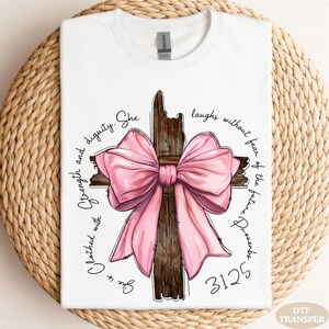 May include: White t-shirt featuring a wooden cross with a large pink bow. The shirt has the text "Clothed with Strength and Dignity. She laughs without fear of the future Proverbs 31:25" in a circular design around the cross.