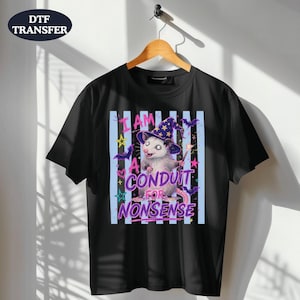 May include: A black short-sleeve t-shirt featuring a graphic of an opossum wearing a purple wizard hat with yellow stars. The text reads I AM A CONDUIT FOR NONSENSE in purple letters. The design includes light blue vertical stripes and bats.