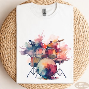 May include: White t-shirt featuring a watercolor design of a drum set with a colorful splash effect. The drum set includes a bass drum, snare drums, and cymbals. The design incorporates shades of red, blue, and yellow.