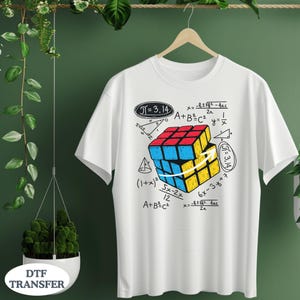 May include: White t-shirt featuring a colorful Rubik's Cube design with mathematical equations and the number Pi. The cube is red, blue, and yellow. The shirt is hanging on a wooden hanger.