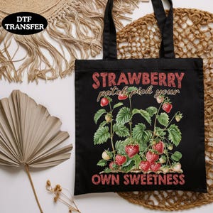 May include: Black tote bag with a strawberry patch design. The bag features a vibrant illustration of strawberry plants with ripe red berries and green leaves. Text on the bag reads "STRAWBERRY patch pick your OWN SWEETNESS."