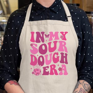 May include: A beige apron featuring the pink and magenta text IN MY SOUR DOUGH ERA in a retro font. The design includes a small butterfly, a disco ball with a crown on top, and tiny flower accents scattered around the bold, wavy lettering.