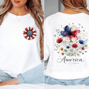 May include: White crew neck t-shirt featuring a patriotic floral design. The front has a sunflower with red, white, and blue petals. The back displays a floral arrangement with the word "America" in script.
