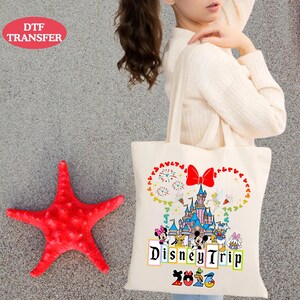 May include: A cream-colored tote bag featuring a colorful Disney design. The design includes a castle, fireworks, and Disney characters. The words "Disney Trip 2023" are printed below the characters. A red starfish is on the left.