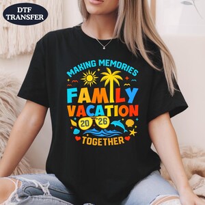 May include: Black t-shirt with a colorful graphic that reads "MAKING MEMORIES FAMILY VACATION 2026 TOGETHER." The design includes a sun, palm tree, waves, and other vacation-themed elements.