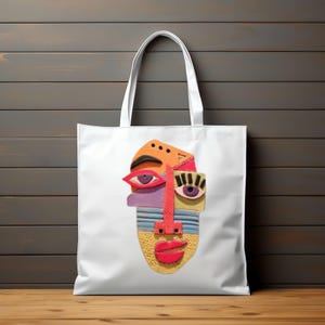 May include: White tote bag featuring a colorful abstract face design. The face has orange, purple, yellow, and red elements, including eyes, a nose, and lips. The bag has white handles and is set against a wooden background.