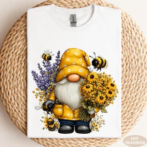 May include: White t-shirt featuring a whimsical gnome design. The gnome is dressed in a yellow outfit and hat, holding a bouquet of yellow flowers. Bees and purple flowers surround the gnome, creating a cheerful, nature-inspired scene.