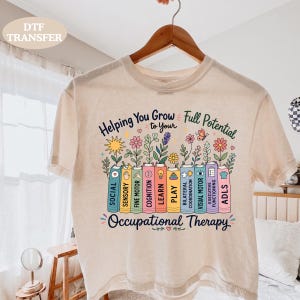 May include: A cream-colored t-shirt featuring a graphic of colorful books with flowers growing from them. The text reads Helping You Grow to Your Full Potential Occupational Therapy, with labels on the book spines including Social, Sensory, Fine Motor, Cognition, Learn, Play, Bilateral Coordination, Visual Motor, Executive Functioning, and ADLs.