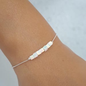 May include: A silver bracelet with a series of small, square, iridescent opal beads. The bracelet is displayed on a wrist, highlighting the subtle colours of the opal stones. The bracelet's chain is thin and elegant, a refined piece of jewellery.