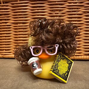 May include: A yellow rubber duck wearing pink glasses and a brown wig, holding a coffee cup and a yellow book with black detailing. The coffee cup has the word "Coffee" on it. The duck is set against a woven basket background.