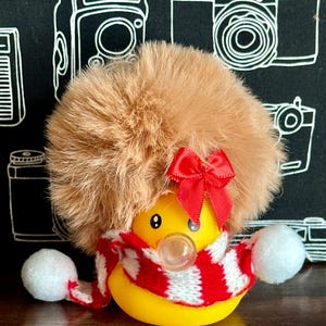 May include: A yellow rubber ducky wearing a fluffy brown hat with a red bow, a red and white striped scarf, and white pom-poms. The background features a black surface with white camera illustrations.