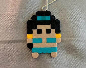 May include: Pixelated keychain featuring a character design with black hair, a teal top, and yellow accents. The keychain is attached to a silver metal keyring. The character is made of small, colorful beads.