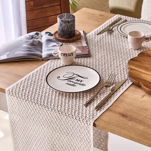 May include: A dining table setting with a cream-colored woven table runner. A white plate with a black rim and the text "My favourite cake" is on the table. A coffee cup, silverware, and a candle are also present.