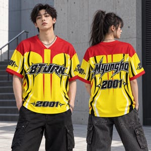 Puede incluir: Una camiseta de manga corta con cuello en V, con una sección superior roja y una inferior amarilla con gráficos negros en forma de llama. El frente muestra el texto 8TURN y 2001, mientras que la parte posterior muestra el texto MYUNGHO y 2001.