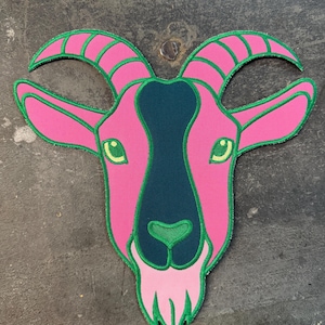 May include: A pink goat head patch with green and teal detailing. The goat features green eyes, horns, and a beard. The patch is designed for application on clothing or accessories.