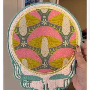 以下が含まれることがあります： ピンクと黄色のキノコ模様の頭蓋骨デザインの刺繍パッチ。 パッチは明るい緑色の縁取りで、衣類やアクセサリーに適しています。