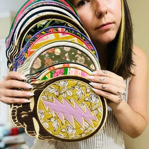 May include: A collection of colorful, round patches with various designs, including floral patterns and a pink lightning bolt. The patches are held up, showcasing their diverse colors and textures. The patches have a brown border.