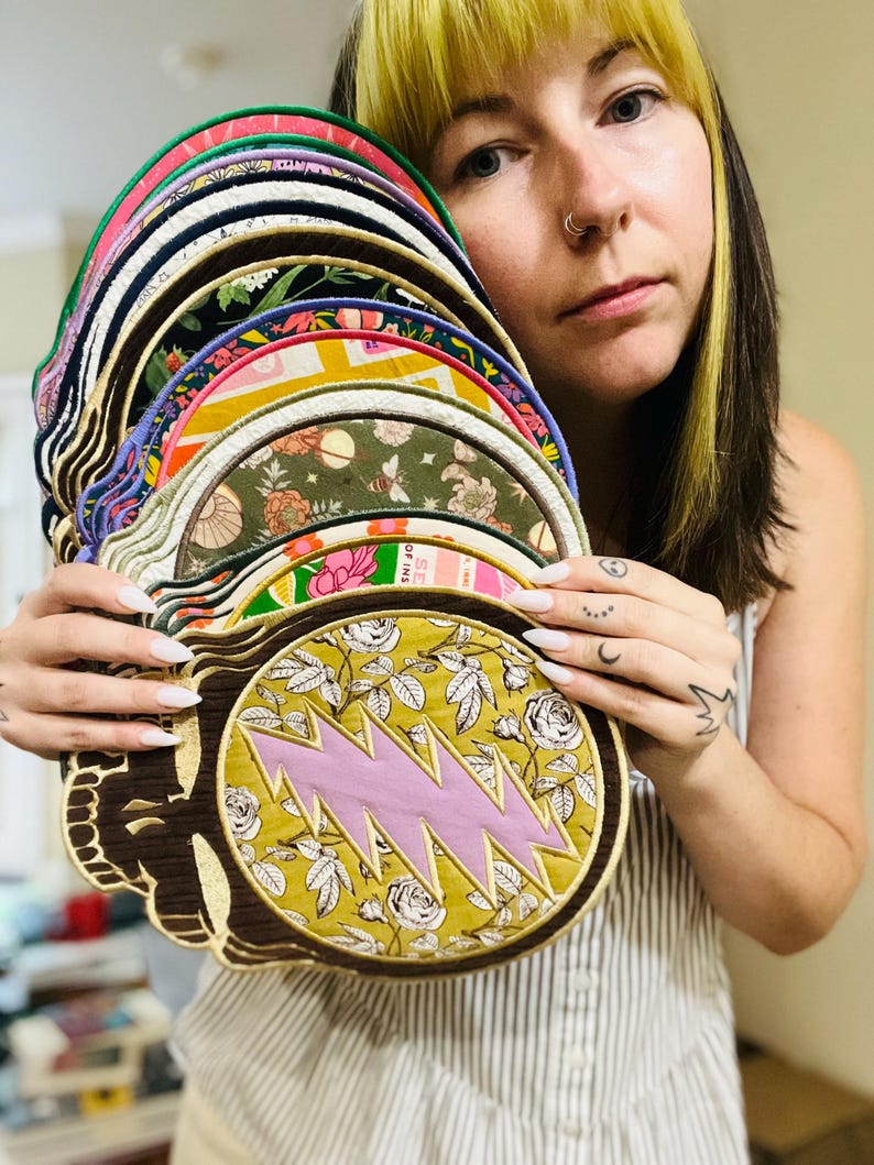 May include: A collection of colorful, round patches with various designs, including floral patterns and a pink lightning bolt. The patches are held up, showcasing their diverse colors and textures. The patches have a brown border.