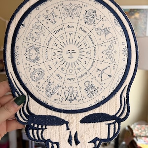 以下が含まれることがあります： クリーム色のパッチ。中央に円形の星座図がある、様式化されたスカルデザイン。図には、海軍ブルーの星座のシンボルと名前が含まれており、海軍ブルーのボーダーが付いています。このパッチはおそらく、衣類やアクセサリー用です。