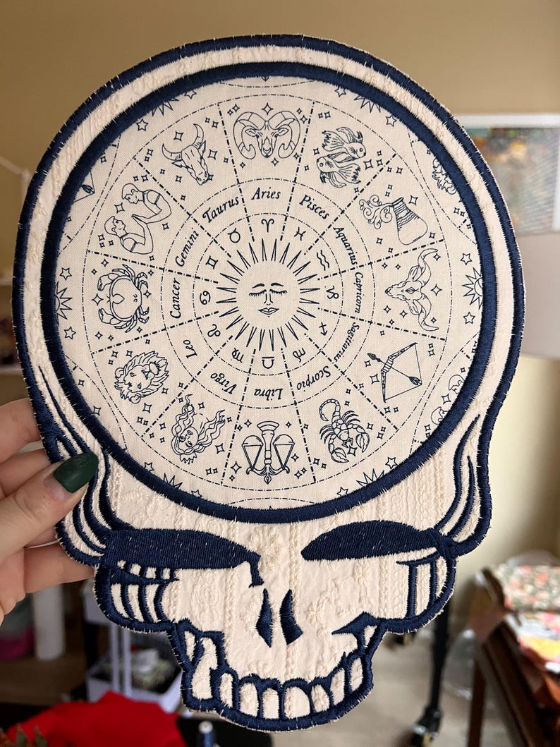 May include: A cream-colored patch featuring a stylized skull design with a circular zodiac chart in the center. The chart includes zodiac symbols and names in navy blue, with a navy blue border. The patch is likely for clothing or accessories.