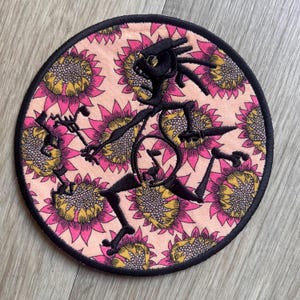 May include: Round embroidered patch featuring a black figure with a stylized design, set against a background of pink and yellow sunflowers. The patch has a black border, creating a defined edge. The design is likely for clothing or accessories.