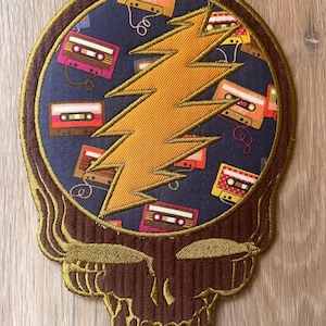 May include: Embroidered patch featuring a skull design with a lightning bolt and cassette tapes. The skull is brown with gold trim, the lightning bolt is orange, and the background is navy blue. The cassette tapes are various colors.