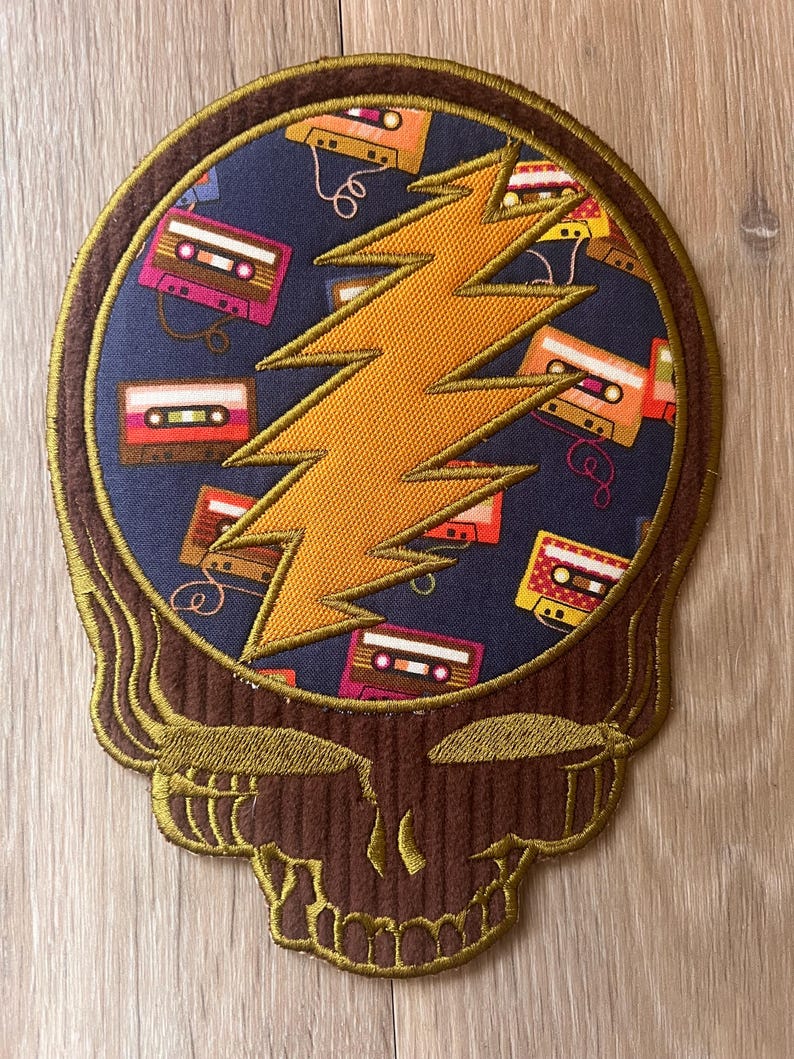 May include: Embroidered patch featuring a skull design with a lightning bolt and cassette tapes. The skull is brown with gold trim, the lightning bolt is orange, and the background is navy blue. The cassette tapes are various colors.