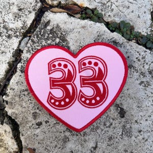 Può includere: Una toppa rosa a forma di cuore con bordo rosso e il numero "33" in rosso. Il numero ha un design decorativo con puntini. La toppa è probabilmente un'applicazione da stirare o cucire.