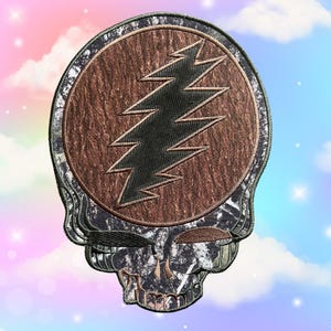 以下が含まれることがあります： 稲妻のエンブレムが付いたスカルデザインの刺繍パッチ。スカルは迷彩柄で、稲妻は黒色です。背景は木目調のテクスチャです。衣類やアクセサリー用です。