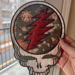 Può includere: Toppa ricamata con un design a teschio, un fulmine rosso e uno sfondo verde con motivi celesti e floreali. La toppa ha un bordo color crema e dettagli del teschio.