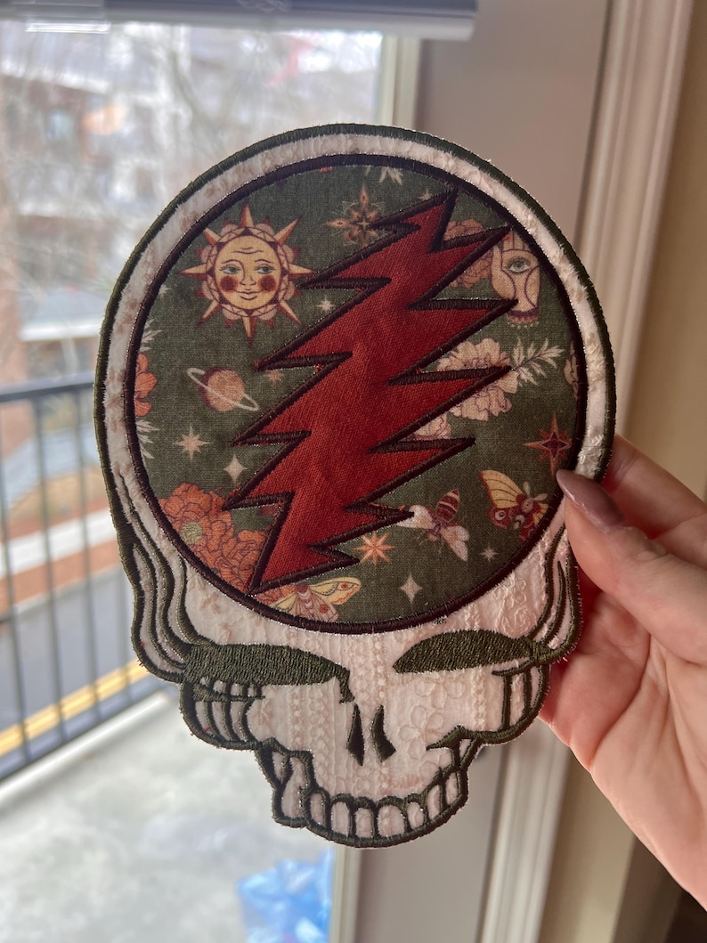 Może przedstawiać: Haftowana naszywka z motywem czaszki, czerwoną błyskawicą i zielonym tłem z niebiańskimi i kwiatowymi wzorami. Naszywka ma kremową obw&oacute;dkę i detale czaszki.